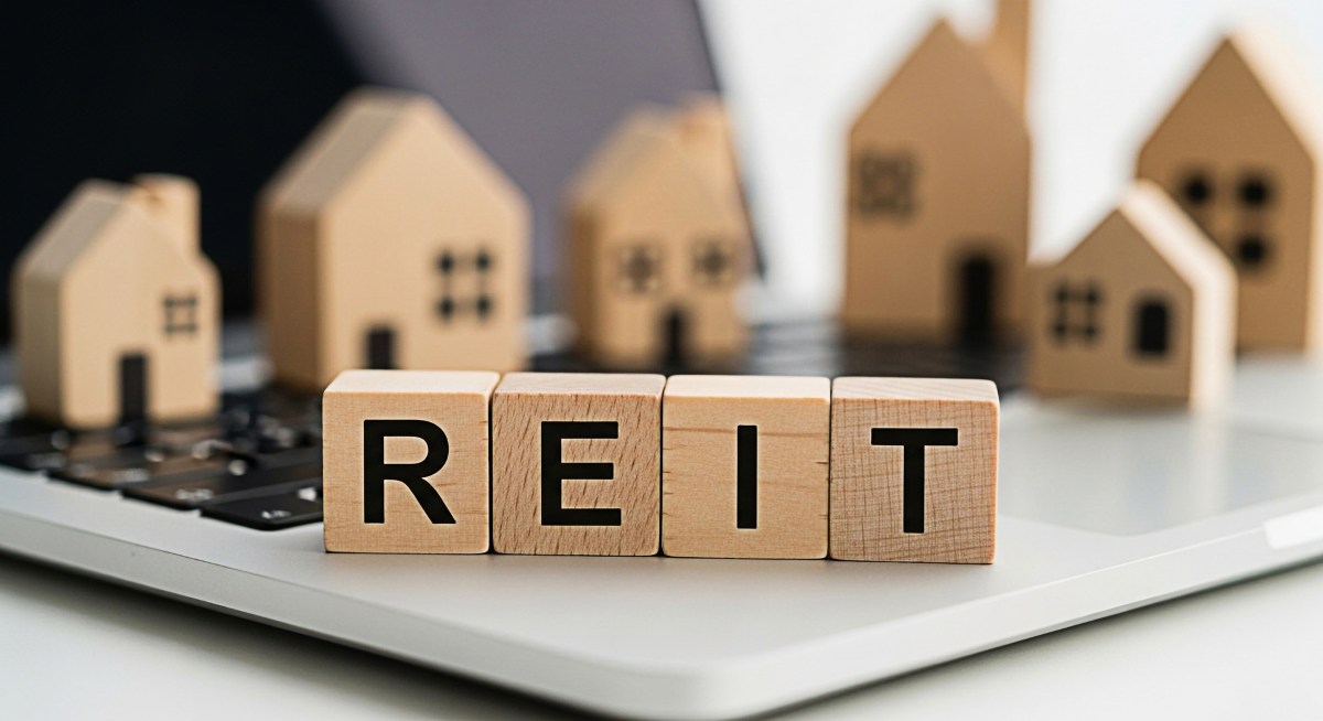 REIT wooden blocks displayed on a laptop keyboard with miniature house models representing real estate investment trusts and financial planning