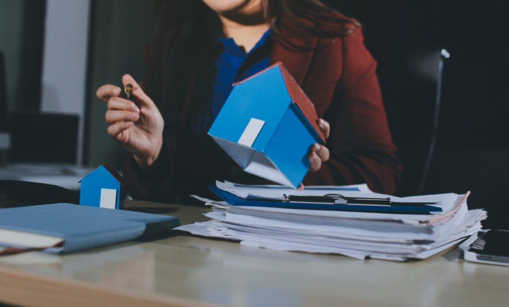 A woman is holding a house model in front of a pile of papers