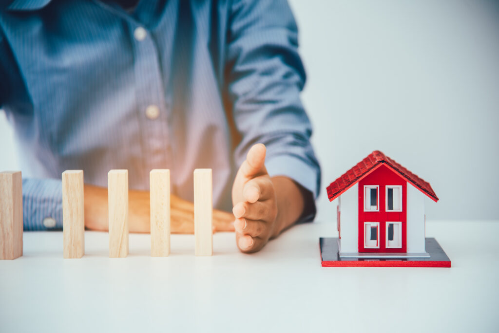 Hand stopping falling wooden blocks from hitting a miniature house, representing risk prevention and property protection