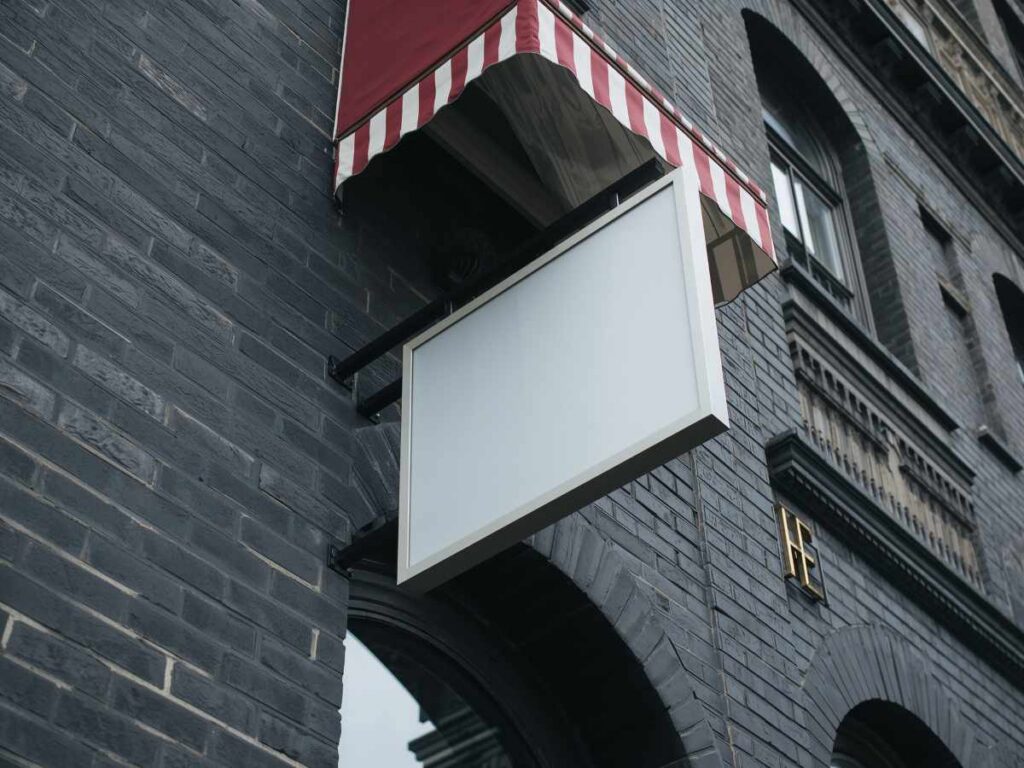 Blank horizontal hanging sign mockup on a dark brick building. White frame sign for a shop
