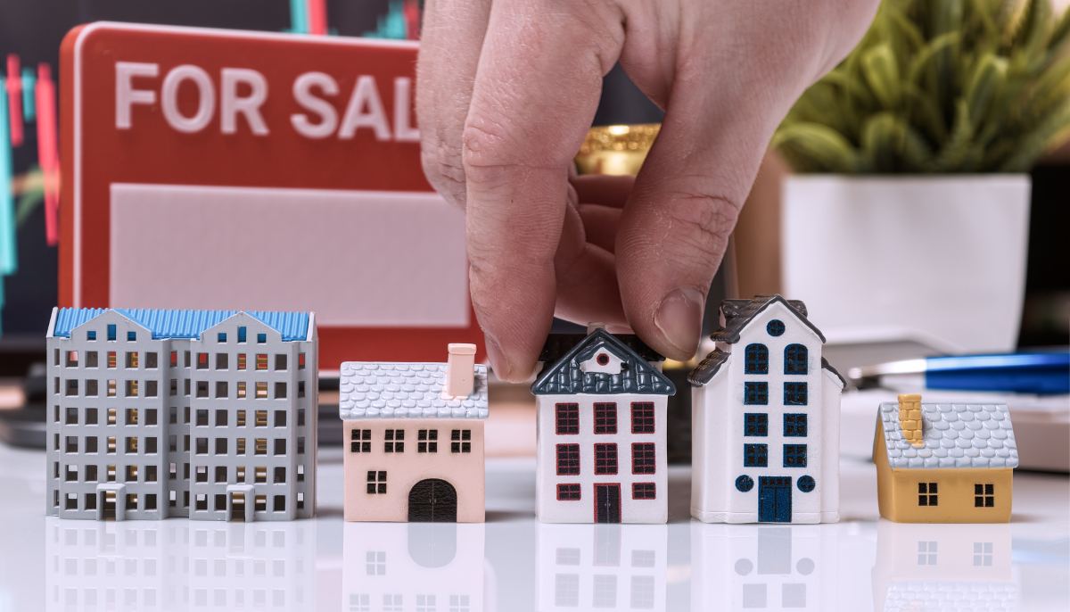 A person holding a miniature house on a desk with a for sale sign