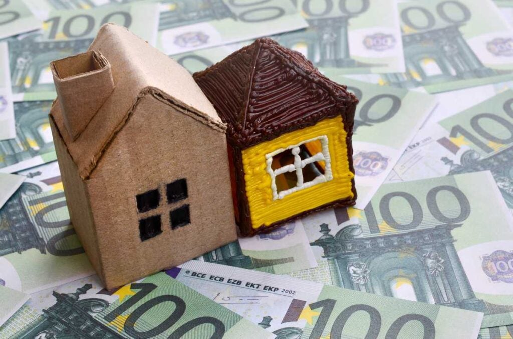 Small toy house is lies on a set of green monetary denominations of 100 euros